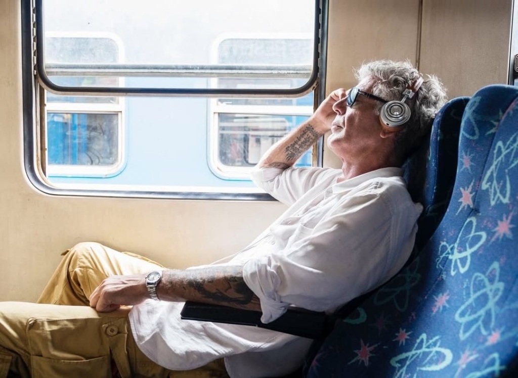 Anthony Bourdain leaning back in a train seat by the window, in headphones and sunglasses, graded warm gold.